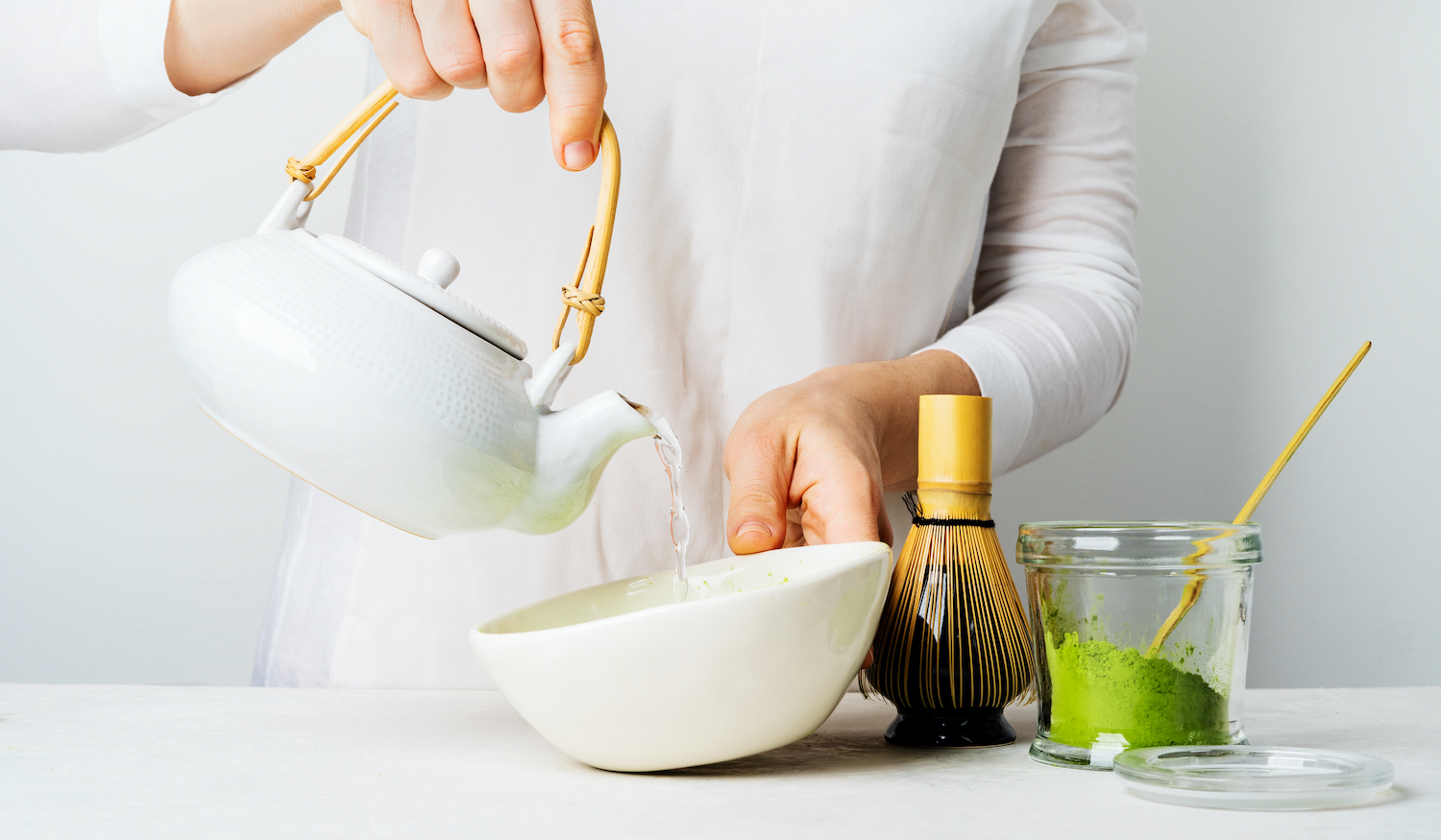 A woman in white pours water from a teapot to make organic Japanese green tea Matcha. Background with tools Chasen bamboo whisk, Chashaku spoon, and bowl for brewing.