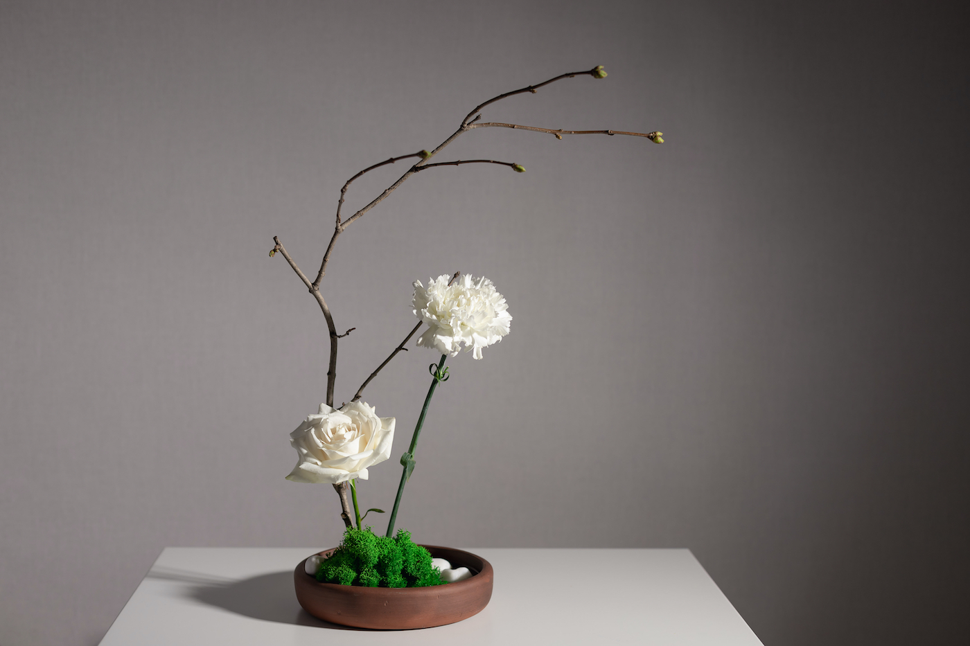 Flower arrangement of white carnation, rose, and tree branches in a ceramic vase with stones and moss. Japanese Ikebana style. gray background