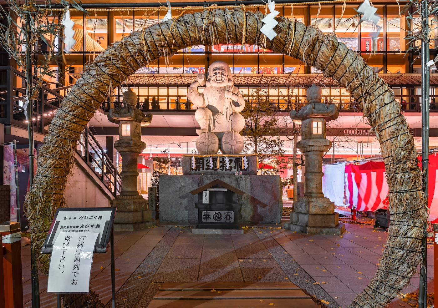 Tokyo, Japan - December 15, 2021: Stone statue of the lucky god Daikokuten illuminated at night during the purification ritual chinowa-kuguri using a circular braided straw gate in the Kanda Myojin shrine.