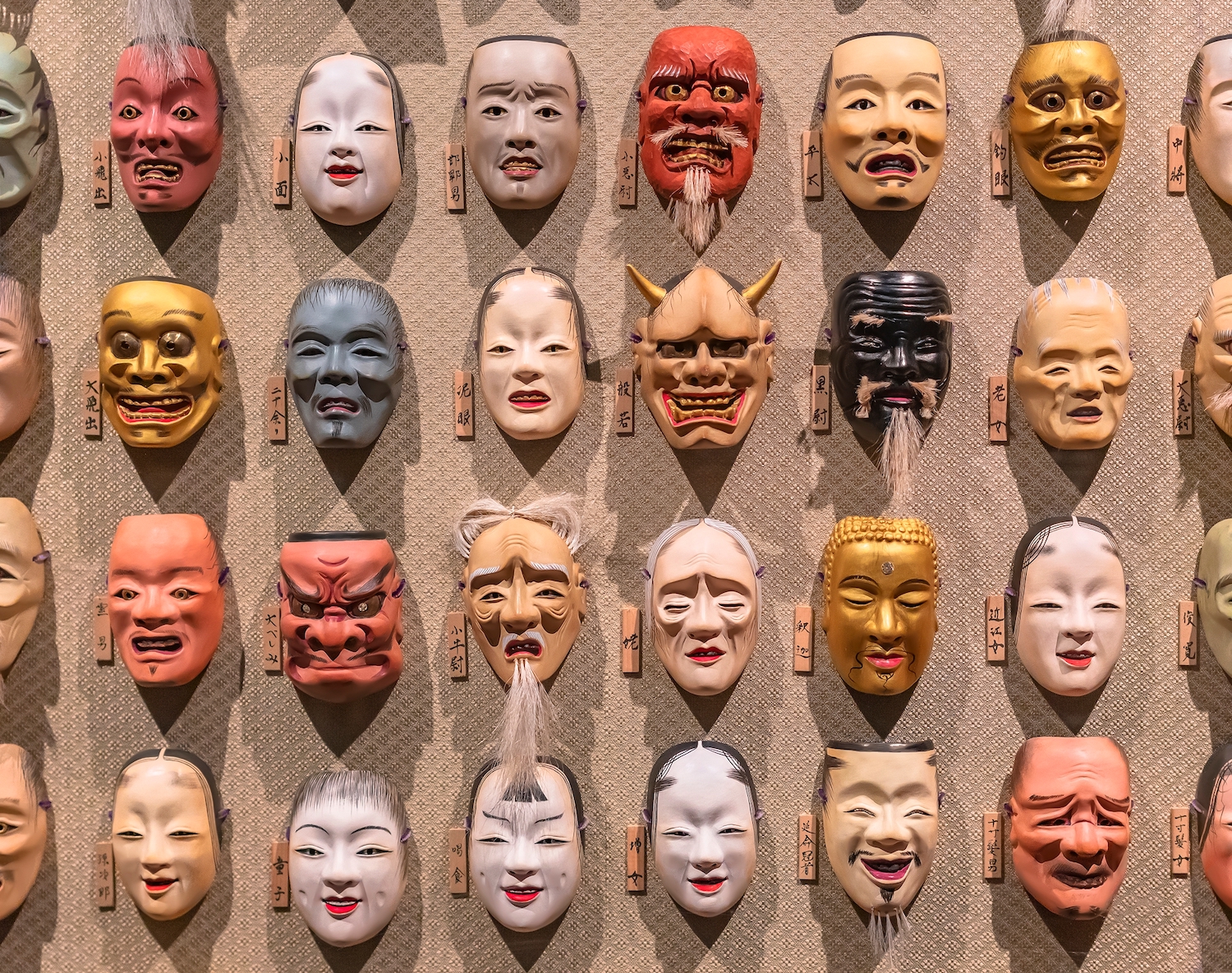 A bunch of Japanese Noh theater masks hanging in rows on a wall depicting various faces expressions belonging to the collection of the ART AQUARIUM Artist Hidetomo Kimura.