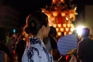 A woman in white and navy Yukata at the festival holding a Uchiwa (fan)